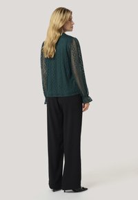 Blusa in pizzo teal con maniche lunghe, texture a fantasia e colletto tondo, abbinata a pantaloni neri a gamba larga e scarpe con tacco nere.