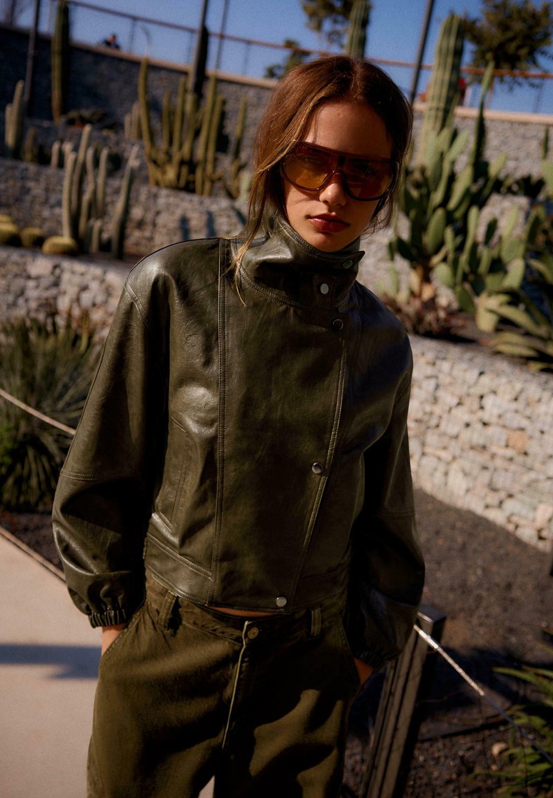 Woman wearing oversized brown sunglasses and olive green leather jacket standing outdoors near cactus plants and stone walls.