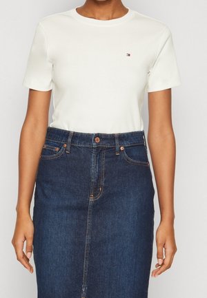 Vrouw draagt een witte nauwsluitende t-shirt met korte mouwen en een hooggesneden donkerblauwe spijkerrok met knoopsluiting aan de voorkant en zakdetails.