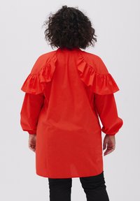 Camicetta di cotone rossa con dettagli arricciati sulle spalle, maniche lunghe e vestibilità comoda, vista da dietro contro uno sfondo neutro.
