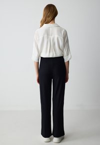 Camicia bianca con maniche arrotolate, abbinata a pantaloni neri a fantasia e sneakers bianche, mostrata da dietro su uno sfondo chiaro.