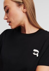 T-shirt noir en coton avec un patch brodé texturé représentant une figure de dessin animé portant des lunettes de soleil sur le côté gauche de la poitrine.