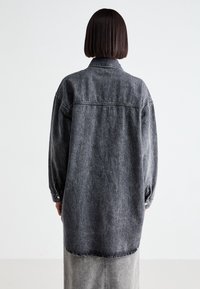 Persona con i capelli scuri e lisci che indossa un lungo giubbotto di jeans neri sbiaditi e una gonna abbinata, in piedi di fronte a un muro bianco.