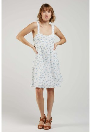 Femme debout, les mains sur les hanches, portant une robe blanche sans manches avec un petit imprimé floral bleu et des sandales marron à brides.