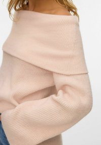 Frau, die einen weichen, hellrosa, schulterfreien Strickpullover mit langen Ärmeln und strukturiertem Rippstoffdetail trägt.