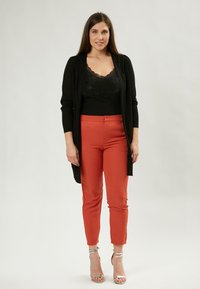 Schwarzer langer Cardigan über einem schwarzen, mit Perlen besetzten Tanktop, kombiniert mit enganliegenden rost-orangefarbenen Hosen und silbernen, geschnürten Absätzen. Glatte Stofftexturen sind sichtbar.