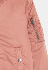 Lyserød bomberjakke med en glat overflade, der har en lynlåslomme på ærmet og en trykknapdetalje foran.