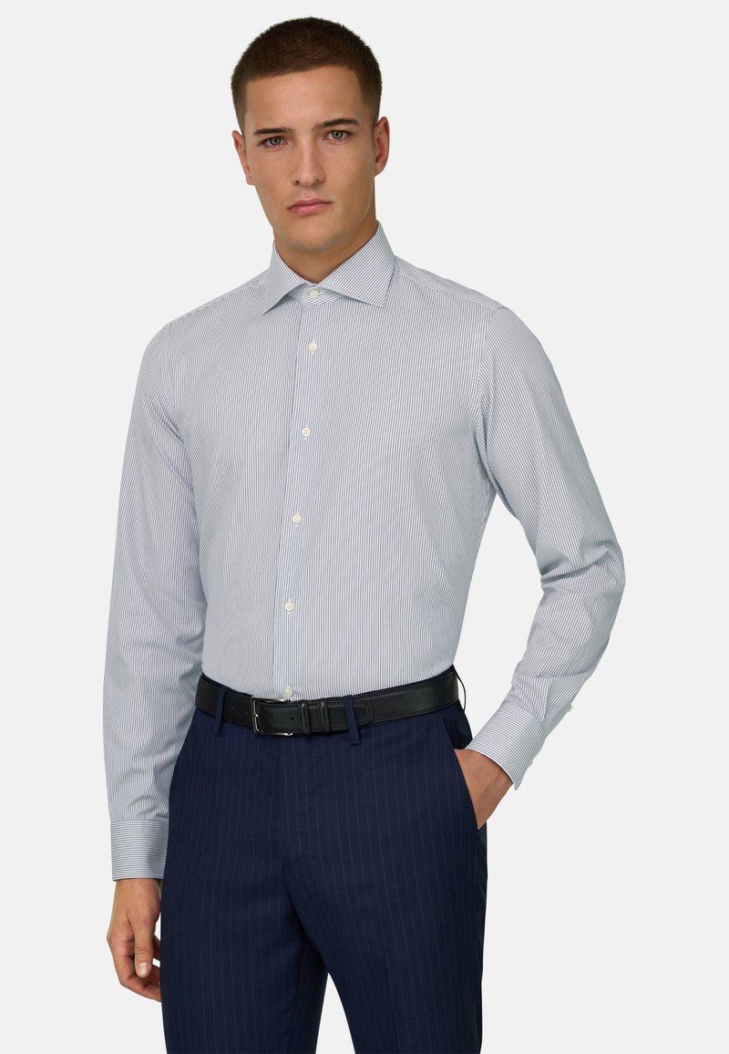 Giovane uomo che indossa una camicia elegante azzurra a righe sottili infilata in pantaloni blu navy a righe sottili con cintura nera, in piedi con una mano in tasca.