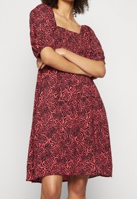 Rotes Kleid mit schwarzem Rosenmuster, kurzen Puffärmelchen, quadratischem Ausschnitt, gerafftem Oberteil und leicht ausgestelltem Rock. Baumwoll-Mischgewebe.