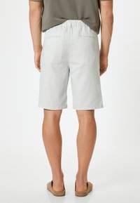 Shorts en lin gris clair avec une taille élastique et deux poches arrière. Finition avec un ourlet roulé, portés avec des sandales à enfiler brunes.