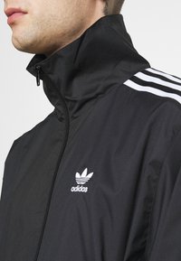 Muž v černé bundě Adidas s vysokým límcem, bílým logem na hrudi a třemi bílými pruhy na rameni.