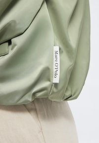 Svijetlozelena tkanina s bijelom oznakom za odjeću ušivenom na šav, na kojoj je ispisan tekst "Marc O'Polo" crnim slovima.