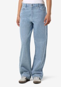 Asmuo dėvi šviesiai mėlynas plačiomis kojinėmis džinsus su mažais cirkonio akmenų papuošimais ir baltus sportinius batelius su juodomis juostelėmis.