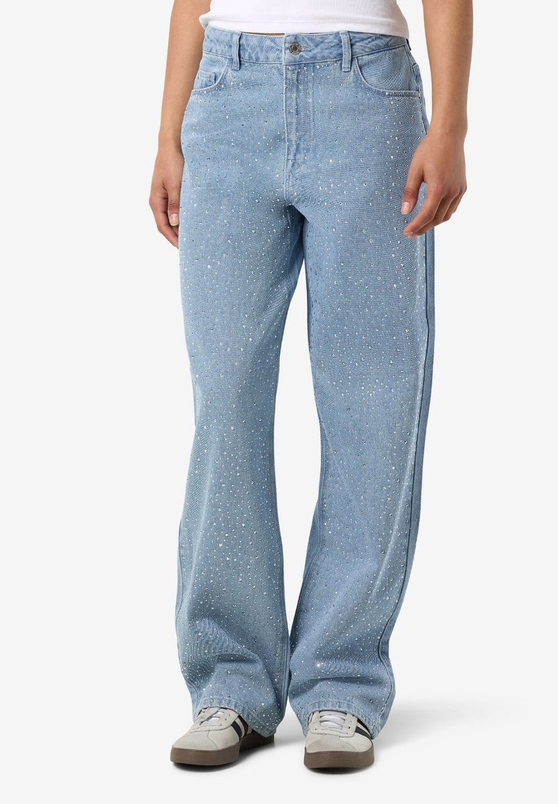 Asmuo dėvi šviesiai mėlynas plačiomis kojinėmis džinsus su mažais cirkonio akmenų papuošimais ir baltus sportinius batelius su juodomis juostelėmis.