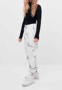 Vita sweatpants med ett svart skorpiontryck, elastisk midja och muddar, bärs tillsammans med en svart ribbad långärmad topp och vita sneakers.