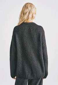 Osoba blondynka o falowanych włosach, ubrana w luźny, ciemnoszary sweter z dzianiny i pasujące ciemne spodnie, odwrócona tyłem na białym tle.