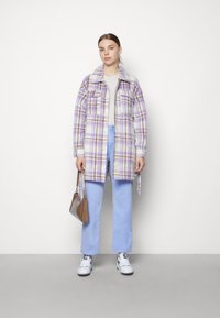 Veste à carreaux violette et blanche avec des accents marron, deux poches poitrine, assortie à un pantalon bleu clair et des baskets blanches. Sac à main marron.