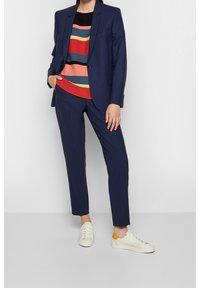 Marinblå kostym med strukturerad kavaj och skräddarsydda byxor, kombinerad med en randig tröja i flera färger och vita sneakers med gula inslag.