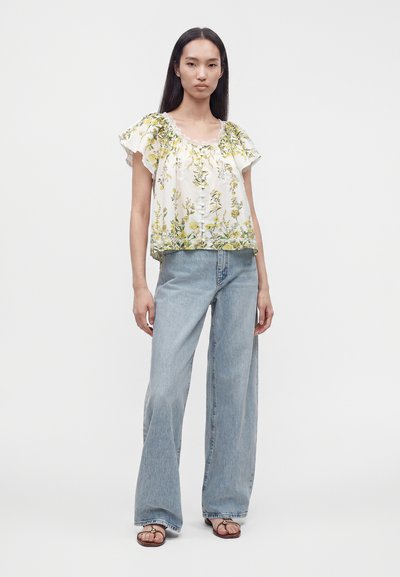 Femme portant un chemisier blanc à manches courtes fleuri, un jean bleu clair à jambes larges et des sandales marron, debout devant un fond uni.