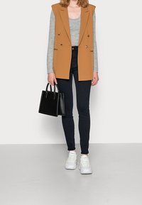 Blazer sin mangas color tostado sobre una camiseta de manga larga gris, combinado con jeans ajustados oscuros y zapatillas blancas, sosteniendo un pequeño bolso negro.