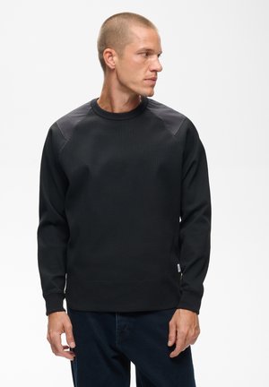 Homme aux cheveux courts portant une chemise noire texturée à manches longues avec des patchs aux épaules, regardant vers la droite sur un fond blanc.