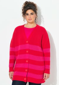 Cardigan rayé rouge et rose avec un décolleté en V plongeant, coupe oversized, fermeture à boutons et texture côtelée. Porté par-dessus un top rose.