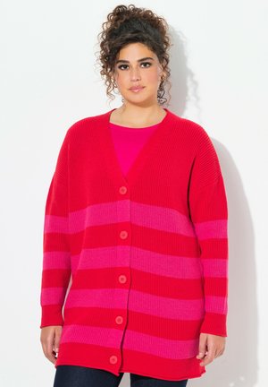 Cardigan rayé rouge et rose avec un décolleté en V plongeant, coupe oversized, fermeture à boutons et texture côtelée. Porté par-dessus un top rose.
