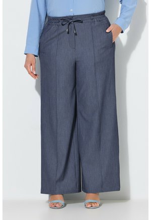 Persoon die donkergrijze wijde broek draagt met een koord in de taille en een lichtblauw overhemd met lange mouwen, staand met één hand in de zak.