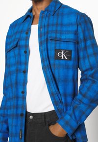 Blue plaid flannel shirt with black accents, button-up design, chest pocket with logo patch, paired with a white T-shirt and black jeans.