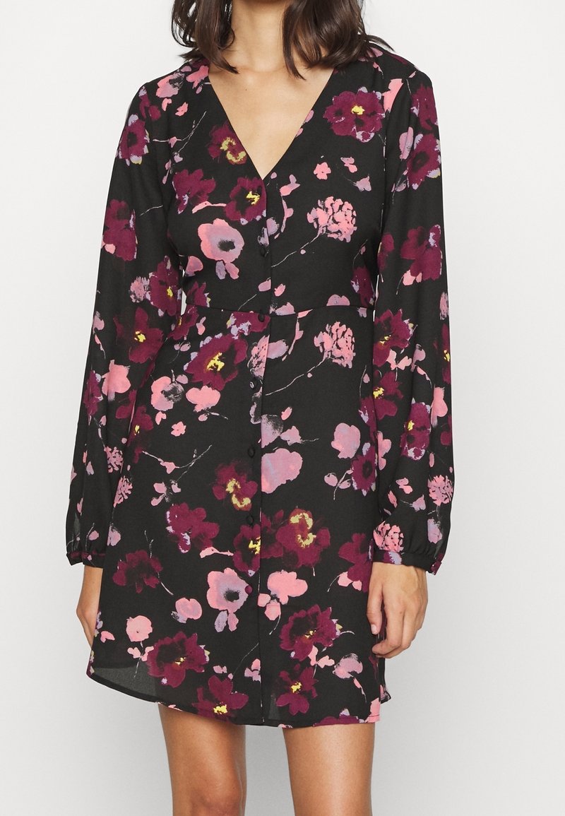 Femme portant une robe noire à manches longues avec des motifs floraux rose, violet et jaune et un décolleté en V, debout devant un fond uni.