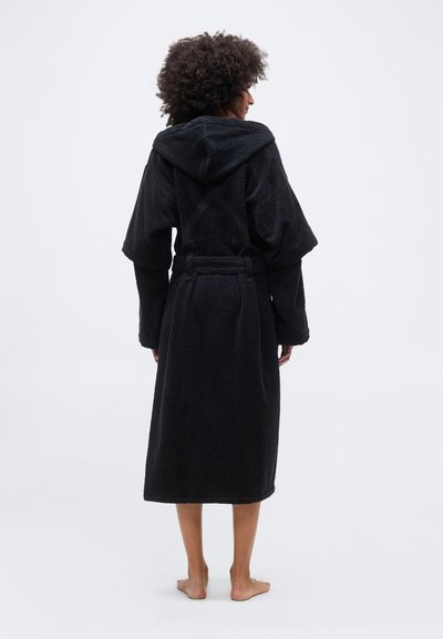 Άτομο με σγουρά μαλλιά στέκεται ξυπόλυτο, φορώντας ένα μακρύ μαύρο ρόμπα με κουκούλα και ζώνη στη μέση, γυρισμένο πλάτη σε ένα απλό φωτεινό φόντο.