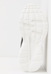 Semelle de basket blanche avec motif de bande de roulement texturé, appuyée à la verticale contre un mur d'angle blanc en arrière-plan.