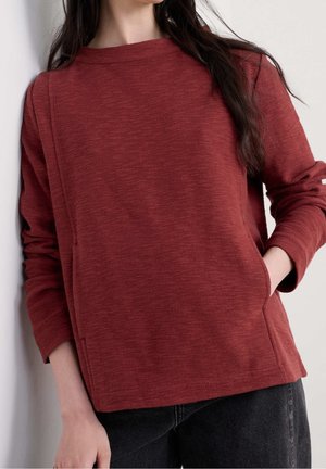 Kvinde iført en langærmet, struktureret, rustfarvet sweatshirt med lommer, kombineret med sorte jeans, stående mod en lys baggrund.