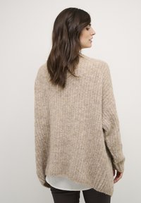 Pull en tricot beige, surdimensionné, avec un tissage texturé et lâche. Comprend des manches longues et un ourlet asymétrique. Porté par-dessus une chemise blanche.