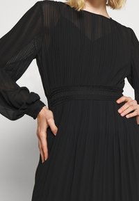 Vestido negro con plisados, mangas largas transparentes, cintura acentuada con encaje y textura de tela fluida. Presenta un escote redondo y bolsillos.