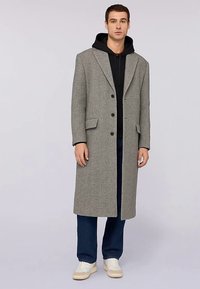 Szary wełniany płaszcz o długim kroju, z otwartymi klapami i trzema guzikami, noszony na czarnej bluzie z kapturem. Zestawiony z ciemnymi dżinsami i białymi trampkami.