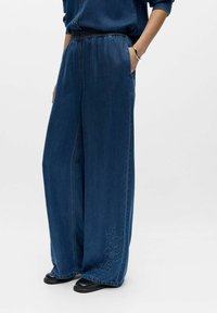 Personne portant un pantalon en denim bleu foncé ample avec une broderie florale près des ourlets, les mains dans les poches, assorti à des chaussures noires et une chemise en denim assortie.