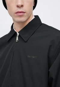 Μαύρο τζάκετ με φερμουάρ και γιακά, με υφή υφάσματος και διακριτικό λογότυπο στο στήθος. Απλός, μινιμαλι στικός σχεδιασμός με μοντέρνα εφαρμογή.