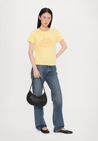Gul bomull T-skjorte med solgrafikk, kombinert med blå slengjeans. Modellen holder en svart vevet håndveske og har på seg svarte leppestifter.