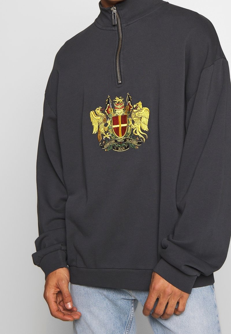 Sweat-shirt noir à quart de fermeture éclair avec un blason héraldique brodé en or et rouge, porté par une personne en jean bleu clair.