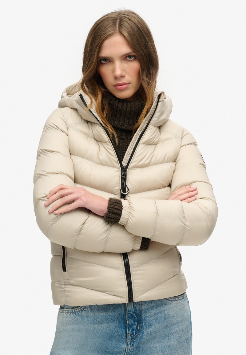 Giacca puff beige con cappuccio, caratterizzata da un design trapuntato, zip frontale e polsini in maglia marrone a coste. Indossata sopra un maglione texturizzato.