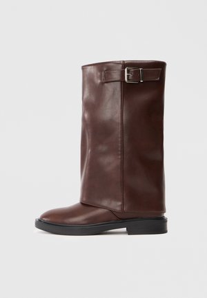Braun lederne kniehohe Stiefel mit glatter Oberfläche, versehen mit einem seitlichen Schnallenakzent und einer runden Zehenpartie, auf einer niedrigen schwarzen Sohle.