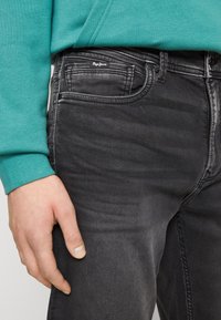 Jeans de mezclilla negros con un ligero desvanecimiento, que presentan un bolsillo frontal, cremallera de metal y un logo de Pepe Jeans en la cinturilla. Mano situada en el costado.