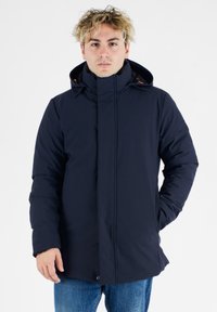 Ung mand med krøllet blond hår iført en marineblå hættetrøje og blå jeans, stående med en hånd i lommen.