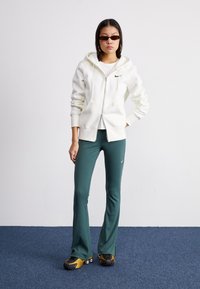 Fato de treino branco com fecho à frente e um logótipo preto, combinado com calças justas verdes. O modelo usa óculos de sol escuros e ténis amarelos e pretos.
