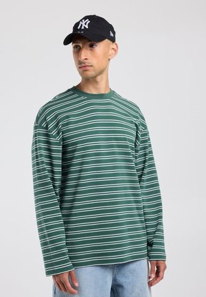 Man draagt een groen-wit gestreept longsleeve shirt, lichtblauwe spijkerbroek en een zwarte baseballpet en kijkt opzij tegen een effen achtergrond.
