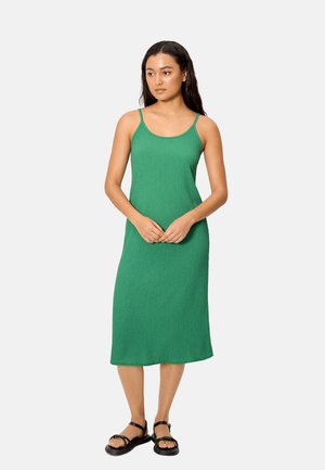 Mujer con un vestido midi verde sin mangas y sandalias negras, de pie con las manos ligeramente entrelazadas, mirando ligeramente hacia la derecha.