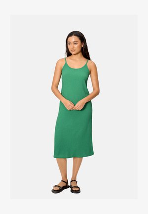 Mujer con un vestido midi verde sin mangas y sandalias negras, de pie con las manos ligeramente entrelazadas, mirando ligeramente hacia la derecha.