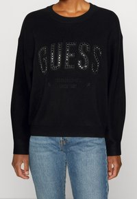 Sweatshirt noir avec logo "GUESS" en paillettes, col rond, manches longues et ourlet côtelé, associé à un jean bleu clair.