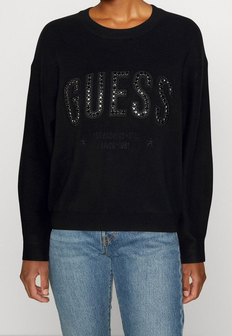 Sweatshirt noir avec logo "GUESS" en paillettes, col rond, manches longues et ourlet côtelé, associé à un jean bleu clair.